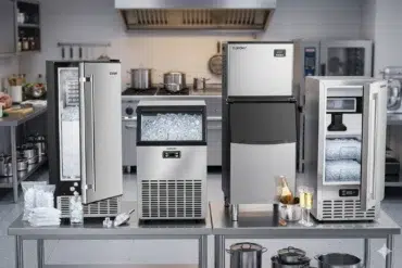 Four stainless steel ice makers on a kitchen counter, filled with ice cubes. The setting is industrial, with a stove and cookware in the background.