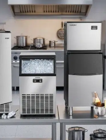 Four stainless steel ice makers on a kitchen counter, filled with ice cubes. The setting is industrial, with a stove and cookware in the background.
