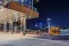 A modern waterfront building with illuminated pillars at night overlooks a cityscape with illuminated boats and skyscrapers.