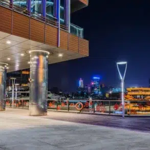 A modern waterfront building with illuminated pillars at night overlooks a cityscape with illuminated boats and skyscrapers.