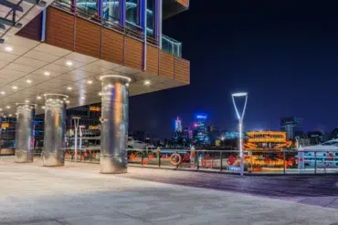 A modern waterfront building with illuminated pillars at night overlooks a cityscape with illuminated boats and skyscrapers.
