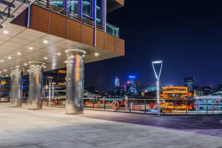 A modern waterfront building with illuminated pillars at night overlooks a cityscape with illuminated boats and skyscrapers.
