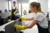 Two office cleaners in uniforms wipe down computers. A woman in the foreground cleans diligently while a man in the background holds a mop, smiling.
