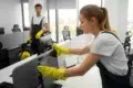 Two office cleaners in uniforms wipe down computers. A woman in the foreground cleans diligently while a man in the background holds a mop, smiling.