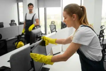 Two office cleaners in uniforms wipe down computers. A woman in the foreground cleans diligently while a man in the background holds a mop, smiling.