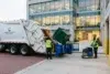 Garbage truck collecting waste outside Axis Professional Center. Two workers in safety vests manage bins, with a modern glass building in the background.