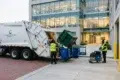 Garbage truck collecting waste outside Axis Professional Center. Two workers in safety vests manage bins, with a modern glass building in the background.