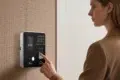 A woman's finger presses the fingerprint scanner on a modern biometric time clock mounted on a textured wall.