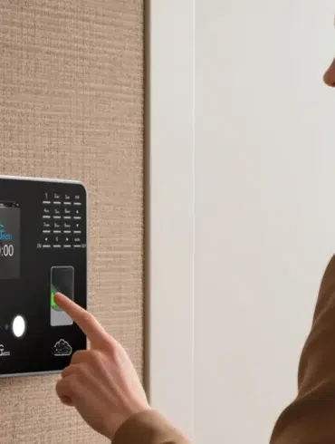 A woman's finger presses the fingerprint scanner on a modern biometric time clock mounted on a textured wall.