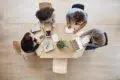 Overhead view of four people collaborating around a light wood table with laptops, tablets, and notebooks.