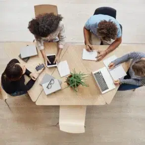 Overhead view of four people collaborating around a light wood table with laptops, tablets, and notebooks.