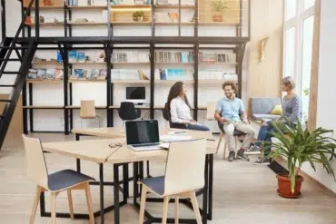 Three people converse in a modern office with bookshelves and collaborative workspaces.