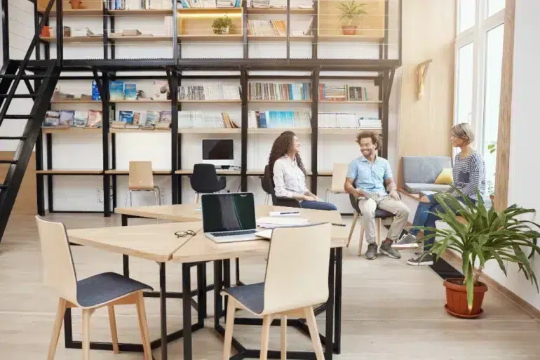 Three people converse in a modern office with bookshelves and collaborative workspaces.