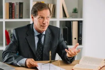 A lawyer in a suit gestures with his hands while sitting at a desk with legal documents, a balance scale, and an open book.