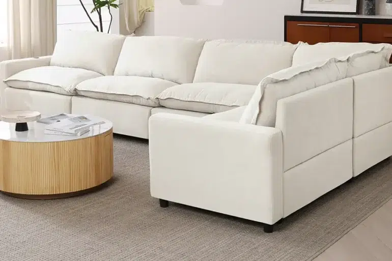 Modern white sectional sofa with plush cushions in a bright living room, featuring a round wooden coffee table on a beige rug.