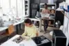 A young woman works on a computer motherboard while a man carries a bin of electronics in a workshop.