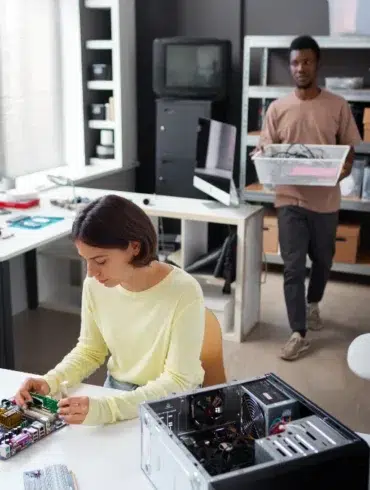 A young woman works on a computer motherboard while a man carries a bin of electronics in a workshop.
