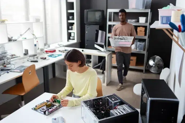 A young woman works on a computer motherboard while a man carries a bin of electronics in a workshop.