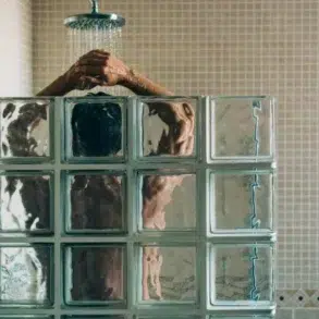 A person stands under a shower with water flowing, partially obscured by frosted glass blocks. The tiled walls add a serene, private atmosphere.