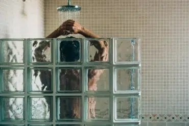 A person stands under a shower with water flowing, partially obscured by frosted glass blocks. The tiled walls add a serene, private atmosphere.