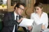 Two professionals in a business setting review documents with a laptop on the table.