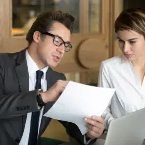 Two professionals in a business setting review documents with a laptop on the table.