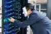 A person in a suit wearing a Guy Fawkes mask interacts with servers in a server room. Blue cables and glowing lights create a mysterious atmosphere.