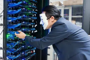 A person in a suit wearing a Guy Fawkes mask interacts with servers in a server room. Blue cables and glowing lights create a mysterious atmosphere.