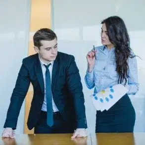 Two business professionals, a man in a suit and a woman with a paper and pen, discuss a project in a modern office.