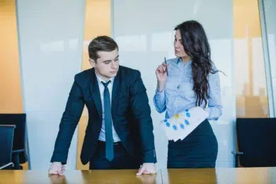 Two business professionals, a man in a suit and a woman with a paper and pen, discuss a project in a modern office.