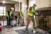 Three construction workers in safety gear and masks renovate an interior. One uses a power tool on a brick fireplace, others clean debris. Bright work lights.