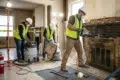 Three construction workers in safety gear and masks renovate an interior. One uses a power tool on a brick fireplace, others clean debris. Bright work lights.