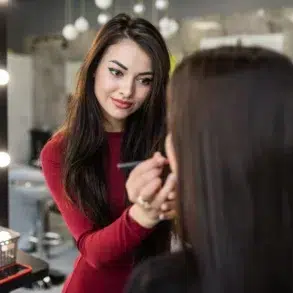 Young woman in a red top applying makeup to a client at a salon. The environment is well-lit, creating a professional and focused atmosphere.
