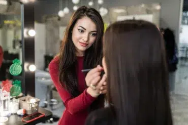 Young woman in a red top applying makeup to a client at a salon. The environment is well-lit, creating a professional and focused atmosphere.