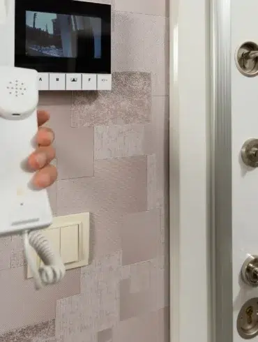 Hand holding an intercom phone in front of a mounted video screen. A secure door with multiple locks is visible, conveying safety and communication.