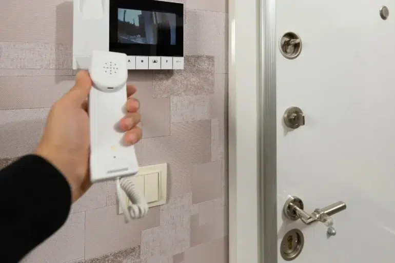 Hand holding an intercom phone in front of a mounted video screen. A secure door with multiple locks is visible, conveying safety and communication.