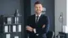 Professional man in a dark suit and tie with arms crossed, standing in a modern office with shelves and a lamp in the background.