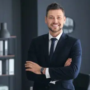 Professional man in a dark suit and tie with arms crossed, standing in a modern office with shelves and a lamp in the background.