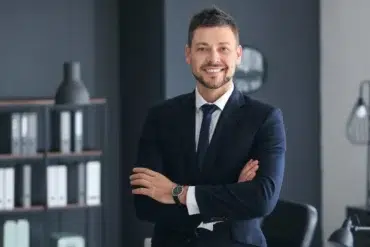 Professional man in a dark suit and tie with arms crossed, standing in a modern office with shelves and a lamp in the background.