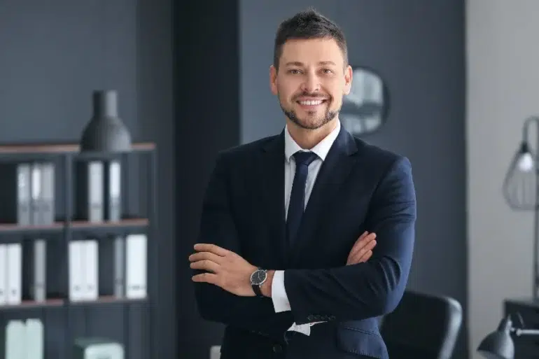 Professional man in a dark suit and tie with arms crossed, standing in a modern office with shelves and a lamp in the background.