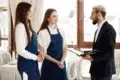 A manager in a suit talks to two smiling waitresses in blue aprons inside a bright, elegant restaurant. They appear engaged and professional.