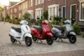 Three scooters in white, red, and silver are parked on a residential street with brick houses and greenery in the background, creating a serene urban scene.