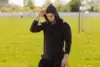 Man in black hoodie adjusting hood while standing in a sunny grassy field with sports goalposts in the background.