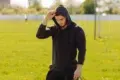 Man in black hoodie adjusting hood while standing in a sunny grassy field with sports goalposts in the background.