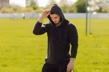 Man in black hoodie adjusting hood while standing in a sunny grassy field with sports goalposts in the background.