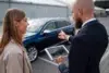 A man in a suit shows a woman a tablet displaying a car while standing near a blue vehicle outdoors, both smiling, suggesting a car sale.