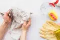 Hands cleaning a glass with a cloth on a white surface. Nearby, a spray bottle, sponge, yellow rubber gloves, and glass cup are neatly arranged.