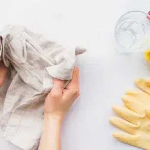 Hands cleaning a glass with a cloth on a white surface. Nearby, a spray bottle, sponge, yellow rubber gloves, and glass cup are neatly arranged.