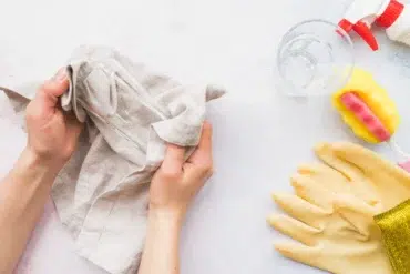 Hands cleaning a glass with a cloth on a white surface. Nearby, a spray bottle, sponge, yellow rubber gloves, and glass cup are neatly arranged.