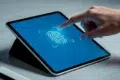 A hand touches a tablet screen displaying a large fingerprint icon on a blue background, symbolizing biometric security. The setting is minimal and modern.